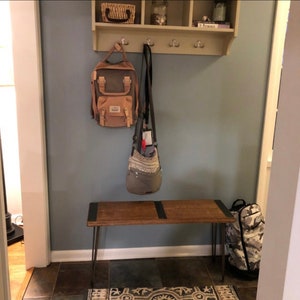 May include: A wooden bench with black hairpin legs sits in a hallway with a shelf above it. The shelf has a basket, jars, and a book on it. There are three hooks on the shelf with a brown backpack and a gray bag hanging from them. A camouflage backpack is on the floor next to the bench.