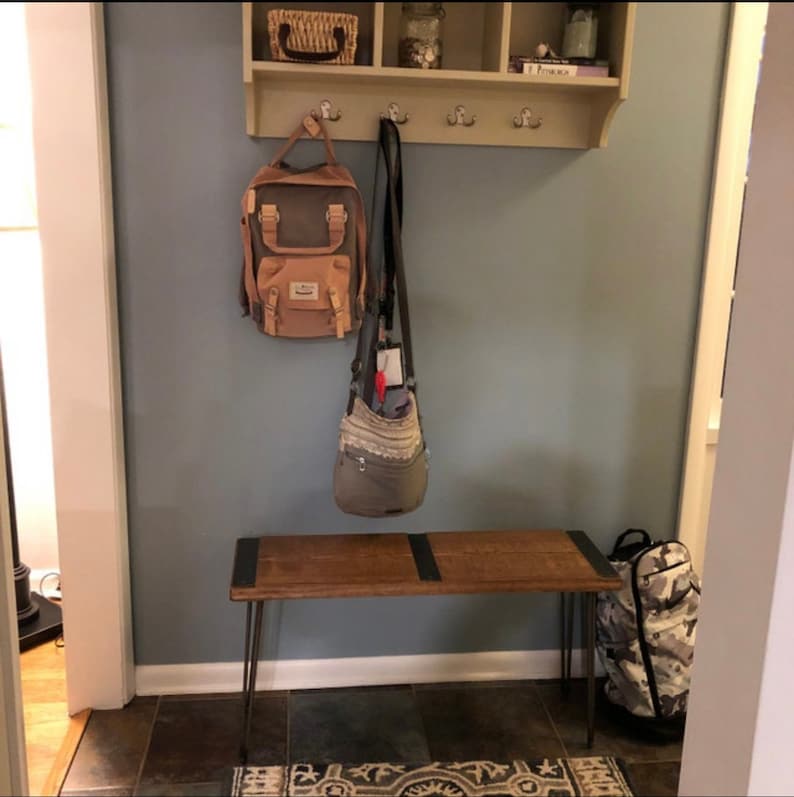 May include: A wooden bench with black hairpin legs sits in a hallway with a shelf above it. The shelf has a basket, jars, and a book on it. There are three hooks on the shelf with a brown backpack and a gray bag hanging from them. A camouflage backpack is on the floor next to the bench.
