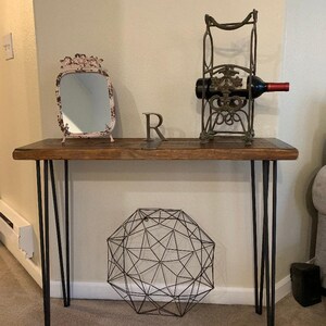 May include: A wooden console table with black hairpin legs. The table has a distressed wood top and is decorated with a small mirror, a wine bottle holder, and a geometric wire orb.