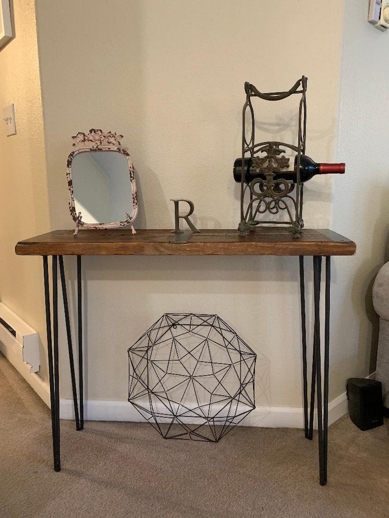 May include: A wooden console table with black hairpin legs. The table has a distressed wood top and is decorated with a small mirror, a wine bottle holder, and a geometric wire orb.