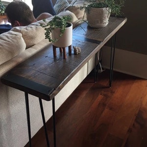 May include: A rustic wooden console table with black metal hairpin legs. The table has a dark wood finish and a black metal trim around the edge. The table is sitting in a living room and has two potted plants on top of it.