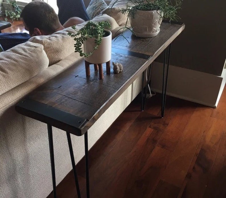 May include: A rustic wooden console table with black metal hairpin legs. The table has a dark wood finish and a black metal trim around the edge. The table is sitting in a living room and has two potted plants on top of it.