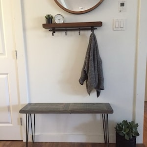 May include: A gray wooden bench with hairpin legs, a gray sweater hanging on a black metal hook rack, a small potted succulent plant, and a clock on a wooden shelf.
