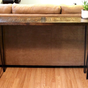 May include: A wooden console table with a dark brown finish and black hairpin legs. The table is in front of a brown leather sofa.