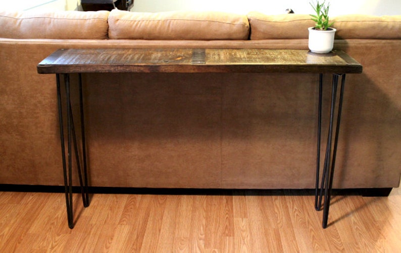 May include: A wooden console table with a dark brown finish and black hairpin legs. The table is in front of a brown leather sofa.