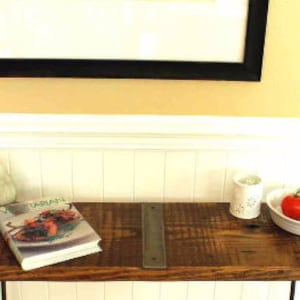 May include: A rustic wooden console table with metal hairpin legs. The table has a distressed wood finish and two metal brackets on the top. A cookbook titled "Vegetarian" is on the table, along with a ceramic candle holder and a bowl of red tomatoes.