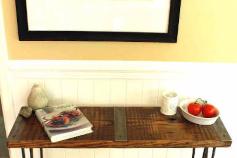 May include: A rustic wooden console table with metal hairpin legs. The table has a distressed wood finish and two metal brackets on the top. A cookbook titled "Vegetarian" is on the table, along with a ceramic candle holder and a bowl of red tomatoes.