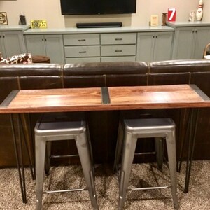 May include: A wooden console table with a rustic brown finish and metal hairpin legs. Two silver metal stools are placed in front of the table. The table is in front of a brown leather sofa.