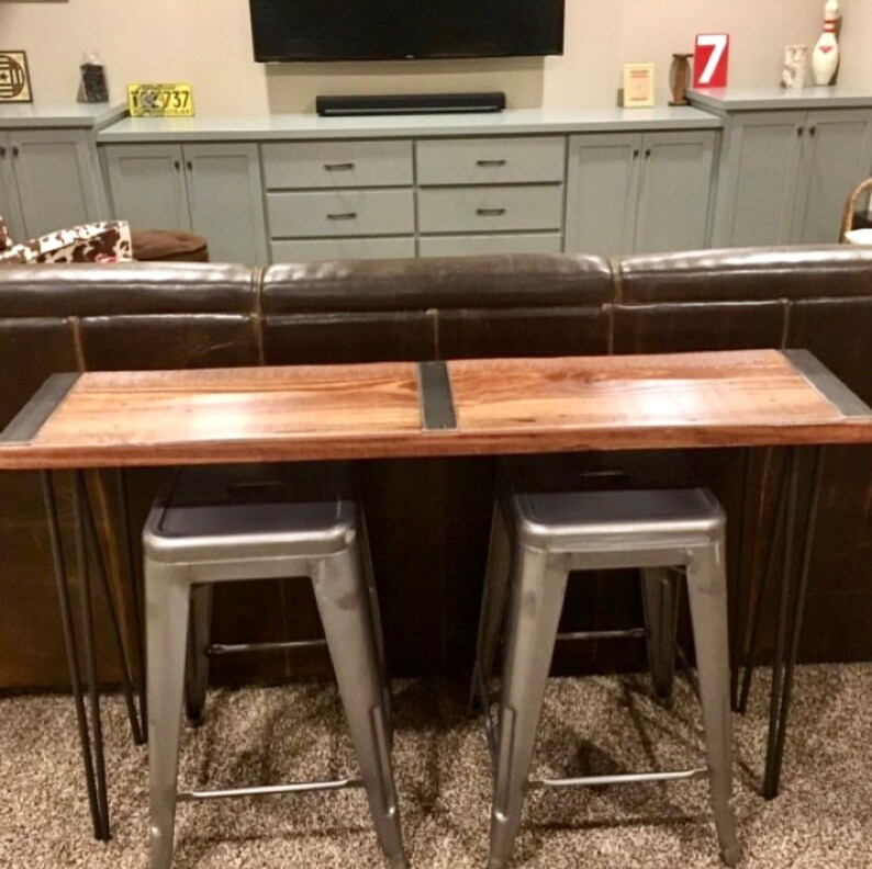 May include: A wooden console table with a rustic brown finish and metal hairpin legs. Two silver metal stools are placed in front of the table. The table is in front of a brown leather sofa.
