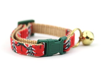 May include: A red cat collar with a Christmas tree pattern, a green buckle, and a gold bell. The collar is made of fabric and has a tan backing. The bell is attached with a gold ring.