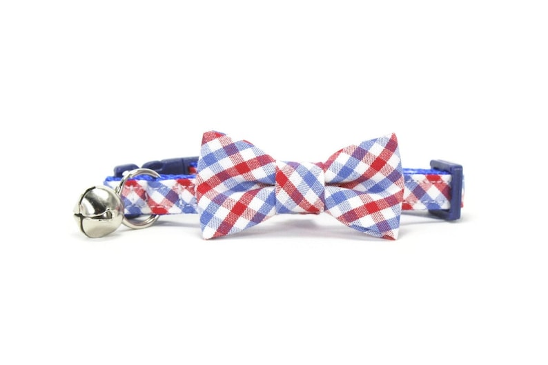 Puede incluir: Un collar de gato azul con una pajarita de cuadros rojos y blancos y una campanilla plateada.