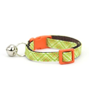Puede incluir: Collar para gato de cuadros verdes y naranjas con una campanilla plateada.