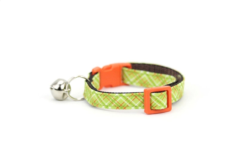 Puede incluir: Collar para gato de cuadros verdes y naranjas con una campanilla plateada.