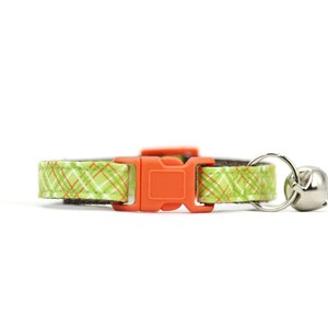 Puede incluir: Un collar para gato de cuadros verdes y naranjas con una hebilla de pl&aacute;stico naranja y una campanilla plateada.