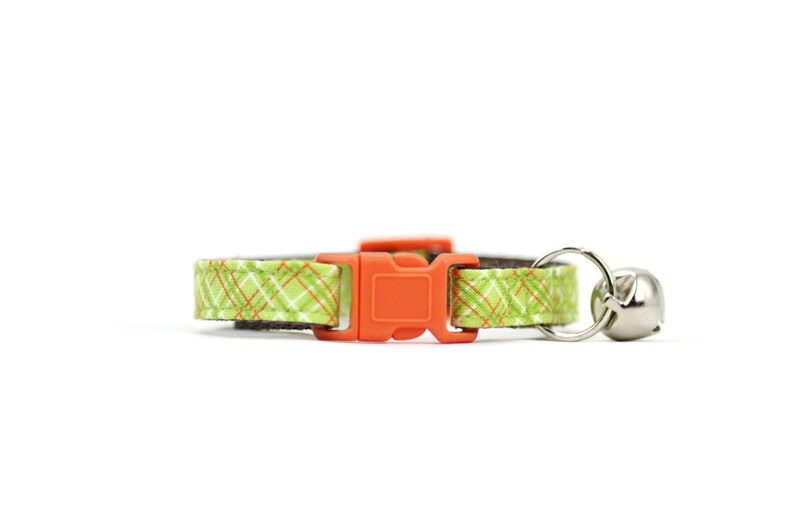 Puede incluir: Un collar para gato de cuadros verdes y naranjas con una hebilla de pl&aacute;stico naranja y una campanilla plateada.