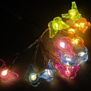 May include: A string of colorful butterfly-shaped LED lights. The butterflies are in shades of pink, blue, green, and orange, with a warm white glow emanating from within. The lights are strung on thin, flexible wires, suitable for decorative use.