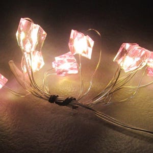 Puede incluir: Una cadena de luces decorativas con formas facetadas de cristal rosa. Las luces están dispuestas en finos cables plateados, con un conector negro. El fondo es oscuro, resaltando los cristales iluminados.