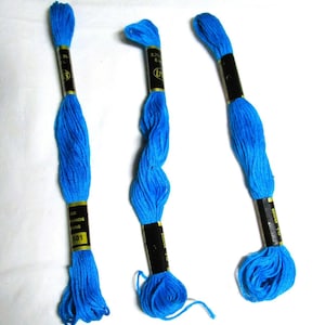 Puede incluir: Tres madejas de hilo de bordar azul brillante. Cada madeja tiene una etiqueta negra y dorada con texto. El hilo está bien enrollado en la parte superior e inferior, con hebras sueltas visibles en la parte inferior. El fondo es blanco.