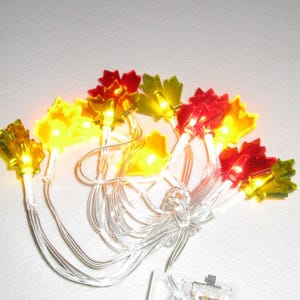 May include: A string of LED lights featuring translucent maple leaf covers in shades of red, orange, and yellow. The lights are connected by a clear plastic wire and include a small battery pack at the base, creating a warm glow when illuminated.