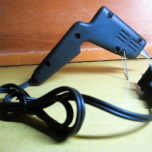 May include: A black hot glue gun with a textured plastic handle and body, featuring a metal wire stand near the nozzle. The tool includes a black power cord with a standard two-prong electrical plug attached at the base of the handle.
