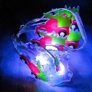 May include: A string of LED lights featuring multiple red truck-shaped ornaments, each carrying a green Christmas tree on its bed. The lights emit a bright white glow, illuminating the translucent plastic figures against a dark background.