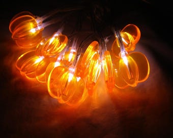 May include: A string of illuminated orange pumpkin-shaped lights. Each translucent plastic pumpkin features vertical ridges and emits a warm, bright glow from an internal light source, connected by a thin wire in a clustered arrangement.