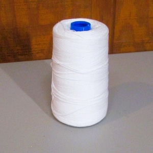 Peut inclure: Une grande bobine cylindrique de fil blanc enroulée étroitement autour d'un noyau central. Le sommet de la bobine présente un capuchon en plastique bleu vif visible au centre de l'enroulement du fil blanc. Le fil a une texture lisse.