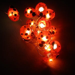 Op de afbeelding: Een lichtsnoer met decoratieve lampen in de vorm van lieveheersbeestjes. De lampen zijn oranje met zwarte stippen. Elke lamp geeft een warm licht, perfect voor feestelijke gelegenheden of voor in huis.