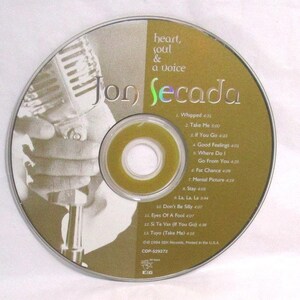 May include: A circular compact disc titled Heart, Soul & a Voice by Jon Secada. The disc features a gold background with a black and white image of a hand holding a vintage microphone. The tracklist includes songs like Whipped and Take Me.