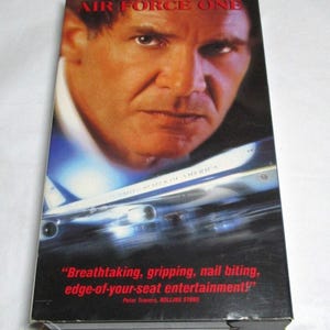 May include: VHS tape for the movie "Air Force One" starring Harrison Ford. The cover features a close-up of Ford's face and an image of the presidential airplane. Text on the cover includes the movie title and a quote from Rolling Stone.