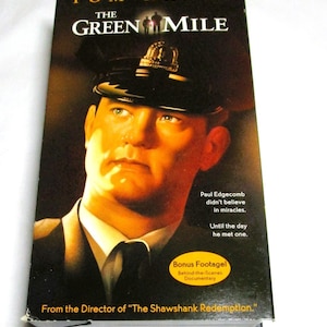 May include: A VHS tape for the movie "The Green Mile" featuring Tom Hanks. The cover has a portrait of Hanks in a police hat. Text includes "Bonus Footage" and "From the Director of 'The Shawshank Redemption.'"