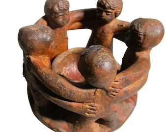 May include: A circular terracotta sculpture featuring four figures standing in a ring, their arms linked around each other. The figures have a textured, reddish-brown finish and surround a central bowl-shaped opening in the middle of the group.