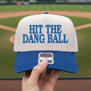 May include: A baseball cap with a white crown and a blue brim. The cap features the embroidered phrase "HIT THE DANG BALL" in blue block letters. The cap is held in front of a baseball field.