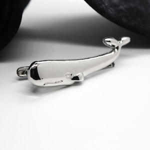 Op de afbeelding: Een gepolijste zilverkleurige metalen dasclip in de vorm van een potvis. Het accessoire heeft een glad, reflecterend oppervlak met een gedetailleerd silhouet van de walvis, inclusief een duidelijk gedefinieerde kop, oog en staartvin.