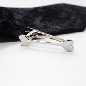 May include: A silver-toned metal tie clip shaped like a dog bone, featuring a spring-loaded hinge mechanism. The tie clip is attached to a black textured fabric necktie, showcasing a polished, reflective surface finish on the bone-shaped ends.