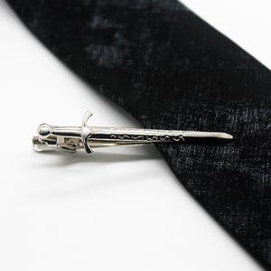 Op de afbeelding: Een zilverkleurige metalen dasclip in de vorm van een zwaard met een gedetailleerd gevest en een recht lemmet, horizontaal geplaatst over een zwarte gestructureerde das. De dasclip heeft een veermechanisme aan de achterzijde voor bevestiging.
