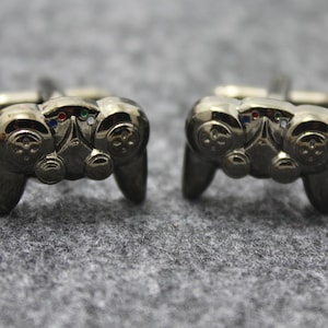 May include: A pair of gunmetal-colored cufflinks shaped like video game controllers. Each cufflink features a detailed design with buttons and directional pads, and small colored accents. The cufflinks are set against a gray, textured background.