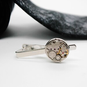 May include: A silver-toned tie clip featuring a circular watch movement mechanism as the centrepiece. The exposed mechanical components include small metallic gears, springs, and tiny red jewels set within a circular frame, attached to a metal bar.