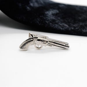 May include: A silver-toned metal tie clip shaped like a revolver handgun. The accessory features a textured grip and a smooth barrel design, with a spring-loaded clip mechanism on the back for securing it to a necktie or other fabric garment.