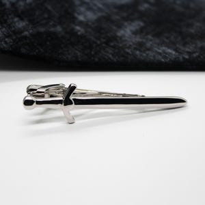 May include: A silver-toned metal tie clip shaped like a sword with a rounded pommel, a crossguard, and a straight blade. The tie clip features a polished, reflective finish and is positioned horizontally against a plain white background.