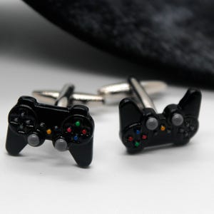 May include: A pair of silver-toned cufflinks featuring black video game controller designs. Each controller has small, colorful buttons in red, green, blue, and yellow, along with two grey analog sticks, attached to a standard metal cufflink backing.