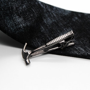 May include: A gunmetal-toned tie clip shaped like a claw hammer with a textured handle, positioned across a black necktie featuring a subtle, mottled fabric pattern. The tie clip has a polished metallic finish and a spring-loaded clasp mechanism.