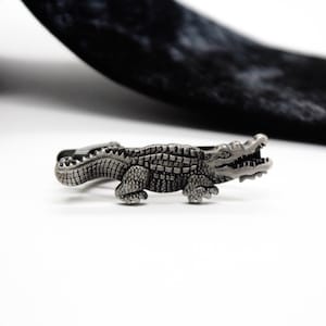 May include: A silver-toned metal tie clip shaped like a detailed crocodile with a textured scaly body, short legs, and an open mouth showing teeth. The crocodile figure is positioned horizontally, featuring a metallic finish with dark accents.