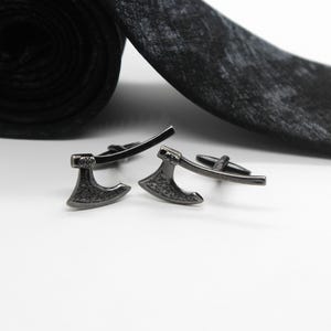 Puede incluir: Un par de gemelos de color gris plomo con forma de hachas de batalla en miniatura, con detalles de hoja texturizados y un diseño de mango curvo, colocados frente a una corbata negra enrollada con un patrón moteado sobre fondo blanco.