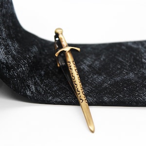 May include: A gold-toned metal tie clip shaped like a sword with an engraved pattern on the blade, positioned on a black necktie featuring a subtle, textured, mottled grey and black fabric pattern throughout the length of the tie.