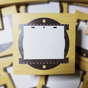 May include: A wooden craft blank with a brown circle cutout and a white rectangle inside. The circle has a ruler design around the edge and two small holes at the top.