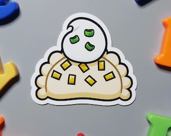 Puede incluir: Una pegatina troquelada con un pierogi de dibujos animados, cubierto con una cucharada de crema agria y rodajas de pimiento verde. El pierogi es de color amarillo claro con relleno cuadrado amarillo. Rodeado de letras magnéticas de colores.