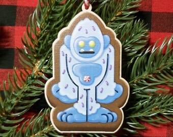 May include: A blue and white Yeti ornament with purple sprinkles and a red and white candy cane. The ornament is made of wood and has a brown background.