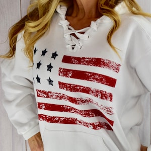 May include: White hooded sweatshirt featuring a distressed American flag design in red and navy blue. The sweatshirt has a lace-up neckline and a front pocket. A casual and patriotic apparel item.