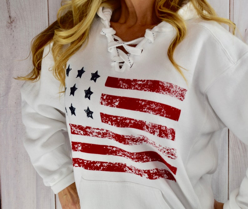 May include: White hooded sweatshirt featuring a distressed American flag design in red and navy blue. The sweatshirt has a lace-up neckline and a front pocket. A casual and patriotic apparel item.